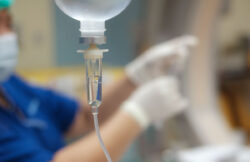 Closeup of an infusion drip, with a blurred background showing nurse's hands preparing another drip.